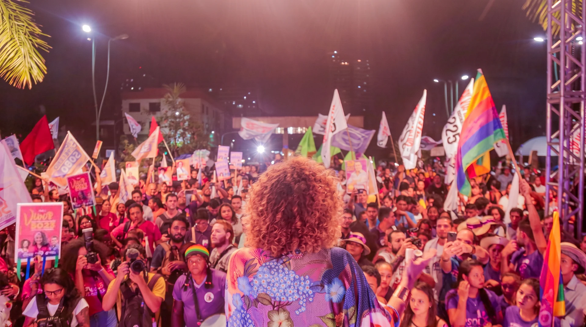 Úrsula vista de costas diante de uma multidão com bandeiras e cartazes em um encontro noturno